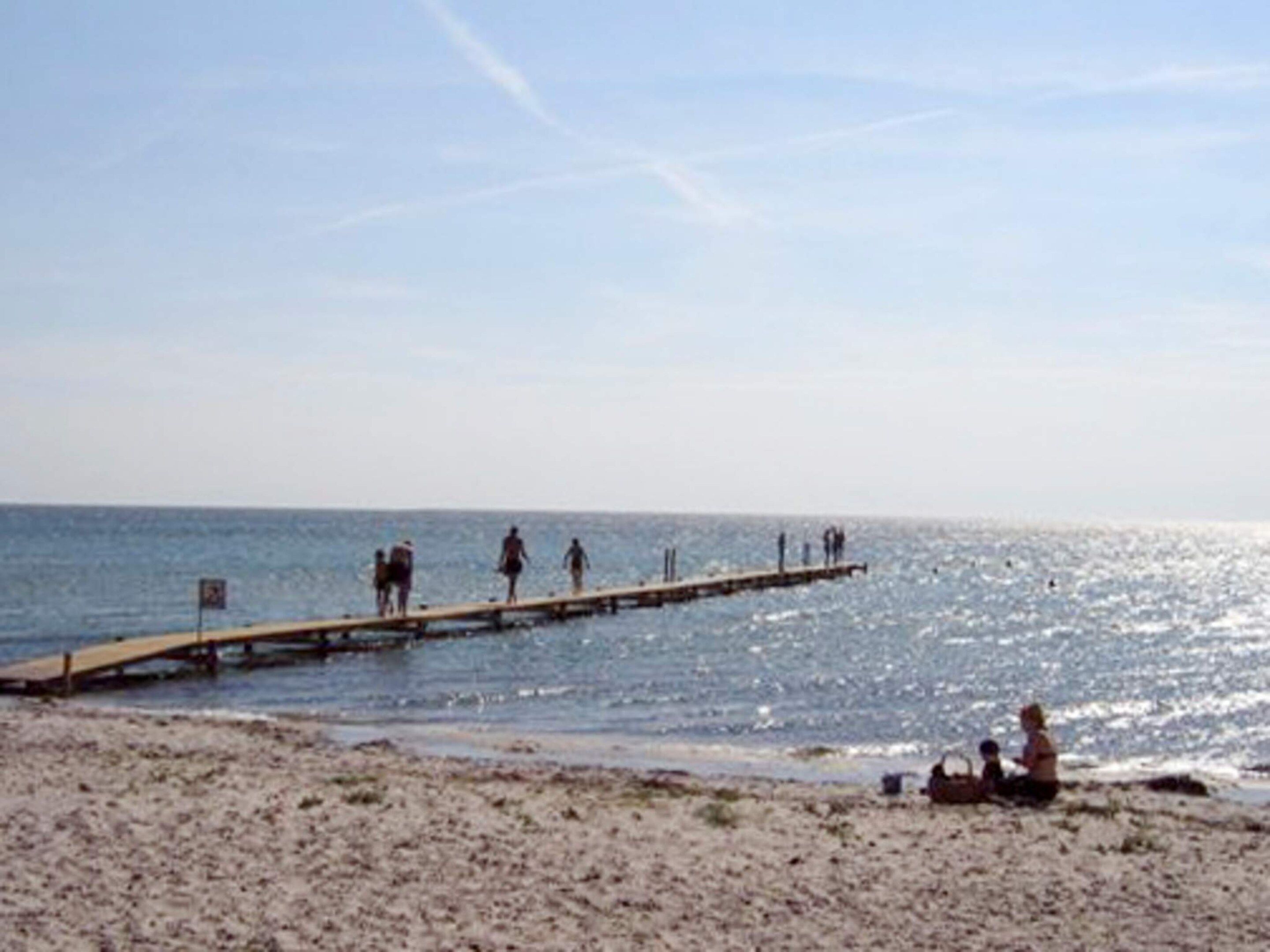 A beach with a wooden pier extending into the water, people walking and sitting on the sand.