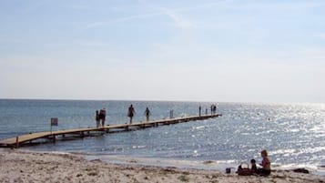 A beach with a wooden pier extending into the water, people walking on it, and a sandy shore with a few individuals sitting.