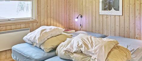 A room with wooden walls, a bed with white and blue bedding, a framed picture on the wall, and a window with a view of trees.