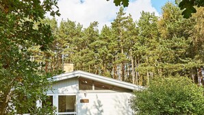 A cabin with a white exterior, surrounded by dense forest, featuring a porch with two chairs and a table. - Cozy Holiday Home in Aakirkeby Bornholm near Sea (Akirkeby)