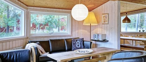A cozy cabin interior with a wooden ceiling, a dining table with benches, and large windows offering a view of trees and a red barn.