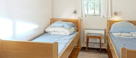 Une pièce avec deux lits en bois, une fenêtre avec des rideaux blancs, un petit tabouret en bois et un radiateur.