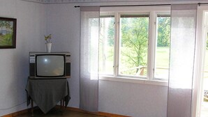 A room with a television on a table, a window with curtains, and a view of greenery outside. - 6 Person Holiday Home in Hacksvik (Håcksvik)
