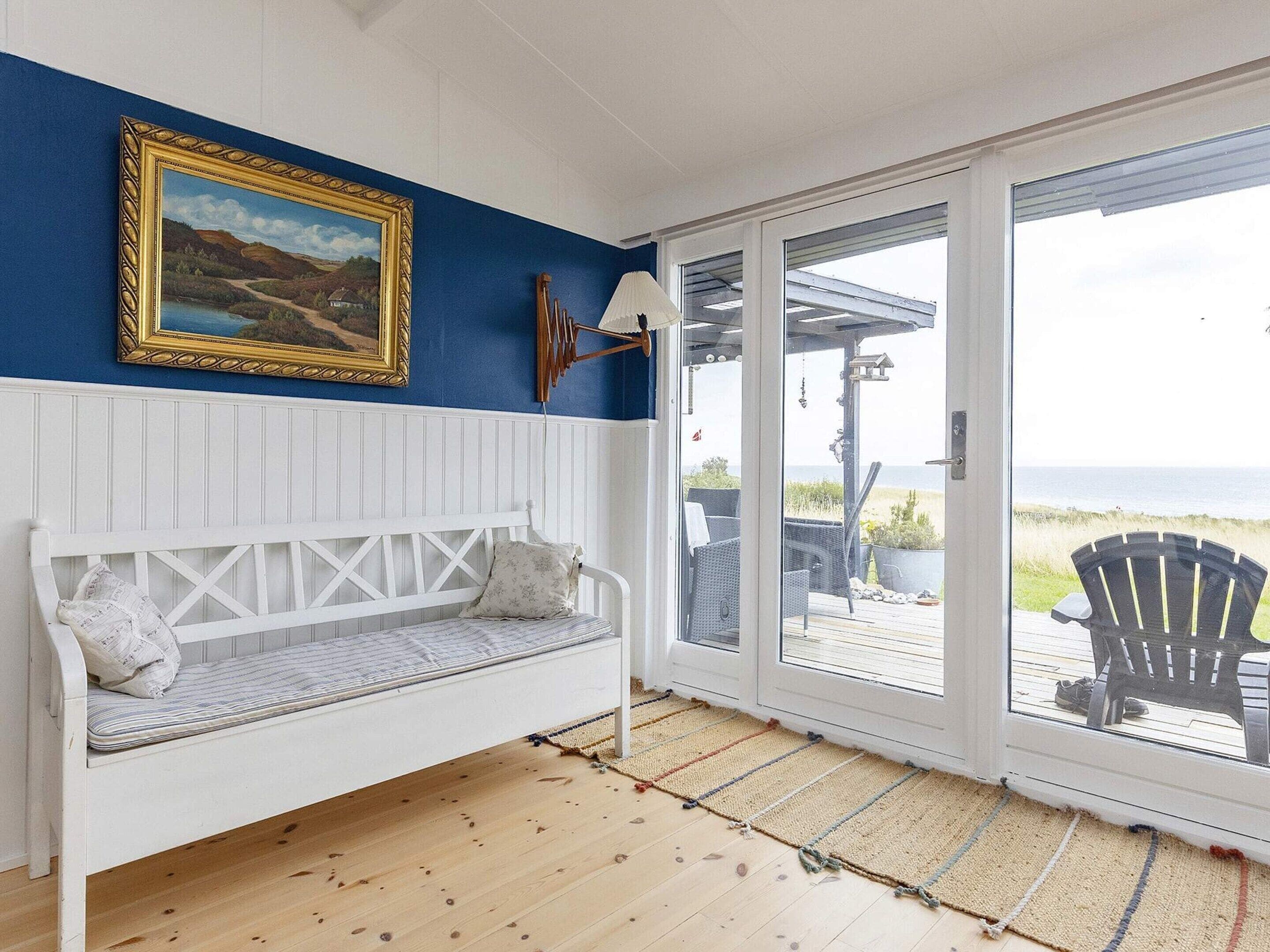 Une pièce avec un banc blanc, un tableau encadré et une vue sur une terrasse extérieure, visible à travers une porte vitrée.