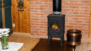 A room with a brick wall, a black wood-burning stove, a copper pot, and a vase with flowers on a wooden table. - 4 Person Holiday Home in Torsby (Torsby)