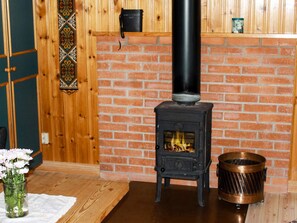 A room with a brick wall, a black wood-burning stove, a copper pot, and a vase with flowers on a wooden table. - 4 Person Holiday Home in Torsby (Torsby)