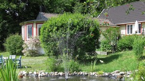 A pond with a fountain, surrounded by a garden with various plants and flowers. There are two houses in the background with red and white trim.