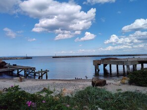 A coastal scene with a wooden pier extending into calm waters, a sandy beach, and a clear sky with clouds.