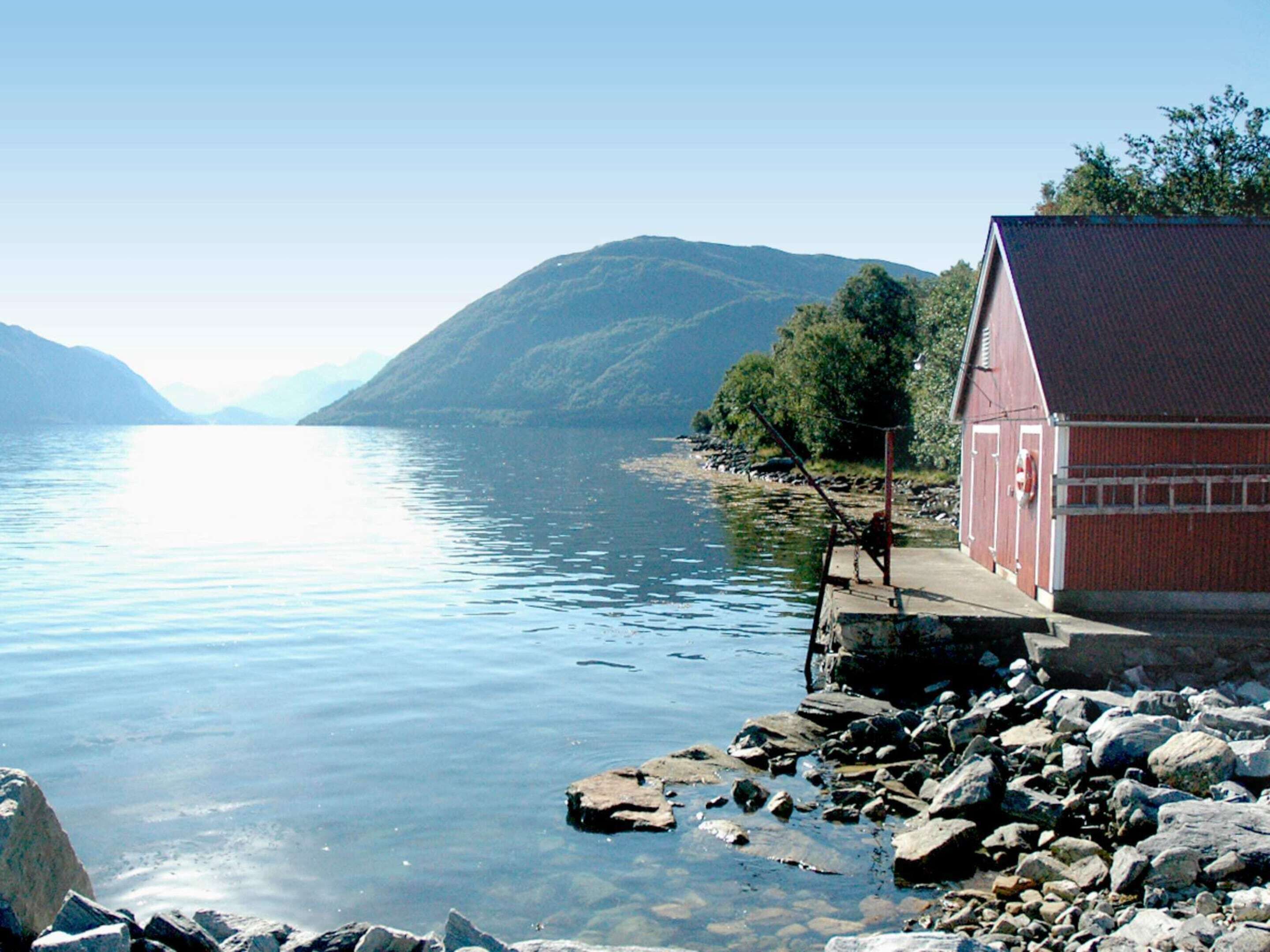A red building with a dock extending into calm water, surrounded by rocky terrain and hills.