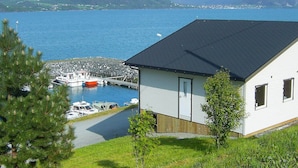 Ein Haus mit dunklem Dach, eine Anlegestelle mit Booten und ein See mit Bergen im Hintergrund.
