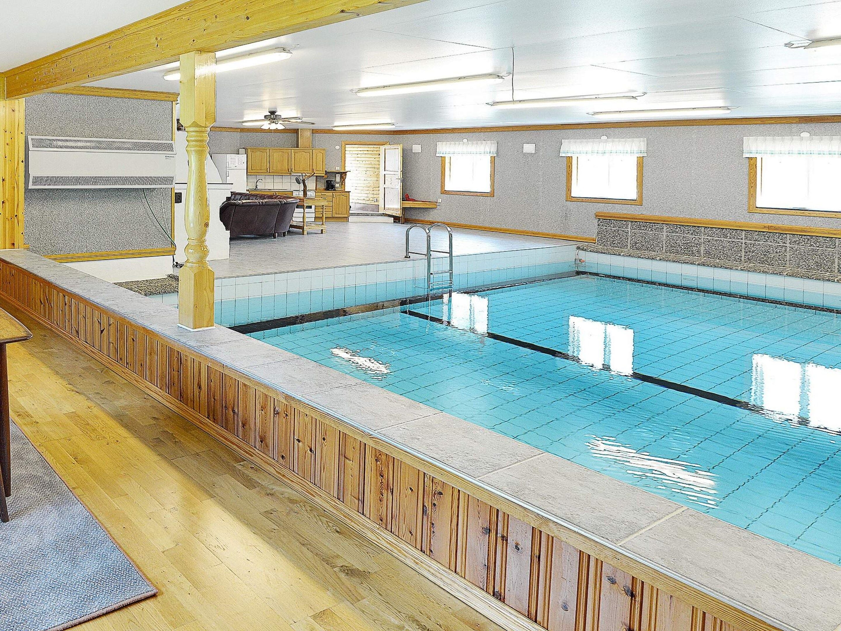 A swimming pool with a wooden deck, a ceiling fan, and a kitchen area in the background.