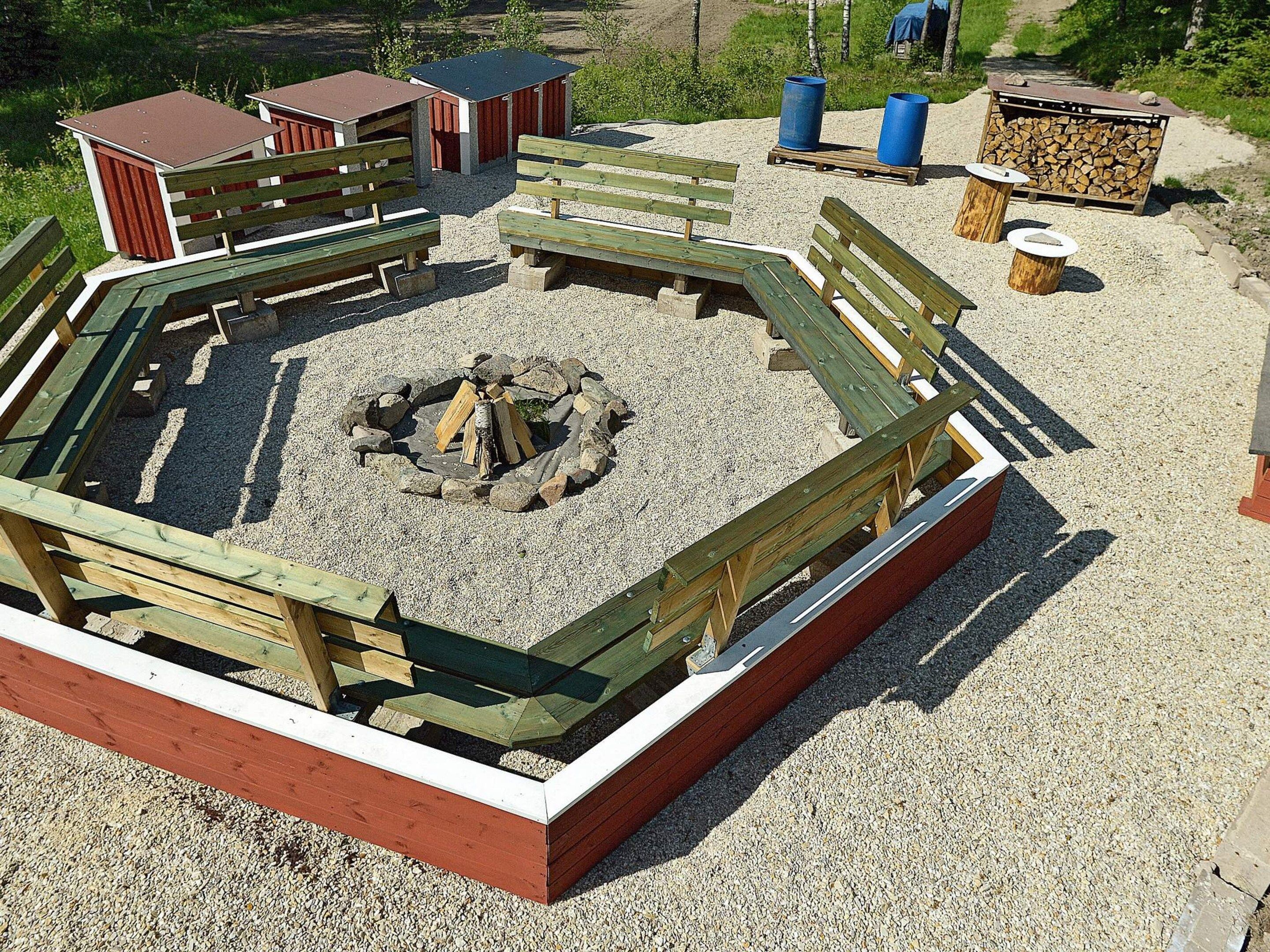 A circular wooden deck with a fire pit in the center, surrounded by benches and a gravel area.