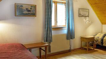 A room with a wooden ceiling, a bed with a red cover, a window with plaid curtains, a radiator, and a small wooden table.