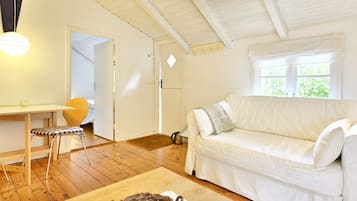 Quarto aconchegante com sofá branco, mesa de jantar com uma cadeira e janela com vista para o verde.