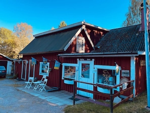 A red building with a blue door and windows, a sign reading "Ingång" and "Entré", and a small outdoor seating area with white chairs. - Holiday Home in Hallabro (Hallar öd)