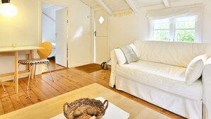 Et hyggeligt rum med en hvid sofa, et spisebord med en stol og et vindue med udsigt til grønne omgivelser.
