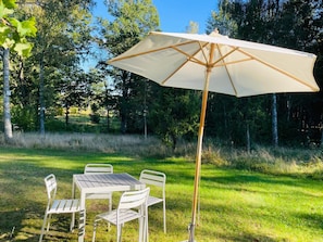 Uma área de refeições ao ar livre com mesa, cadeiras, um grande guarda-sol e um campo gramado com árvores ao fundo.