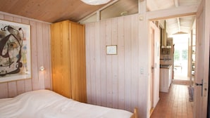 A wooden room with a bed, a wooden wardrobe, and a kitchen visible through an open door.