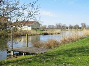 A riverside view with a wooden dock, a house with a red roof, and a grassy bank.