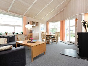 Una sala de estar acogedora con sofá, mesa de centro y comedor con sillas. Hay ventanas con cortinas, una chimenea y una zona de cocina con armarios y estufa.