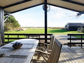 A covered outdoor seating area with a table and chairs, overlooking a grassy field and a distant building.