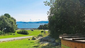 Eine Holzterrasse mit Blick auf eine Wiese, im Hintergrund Meer und Berge.