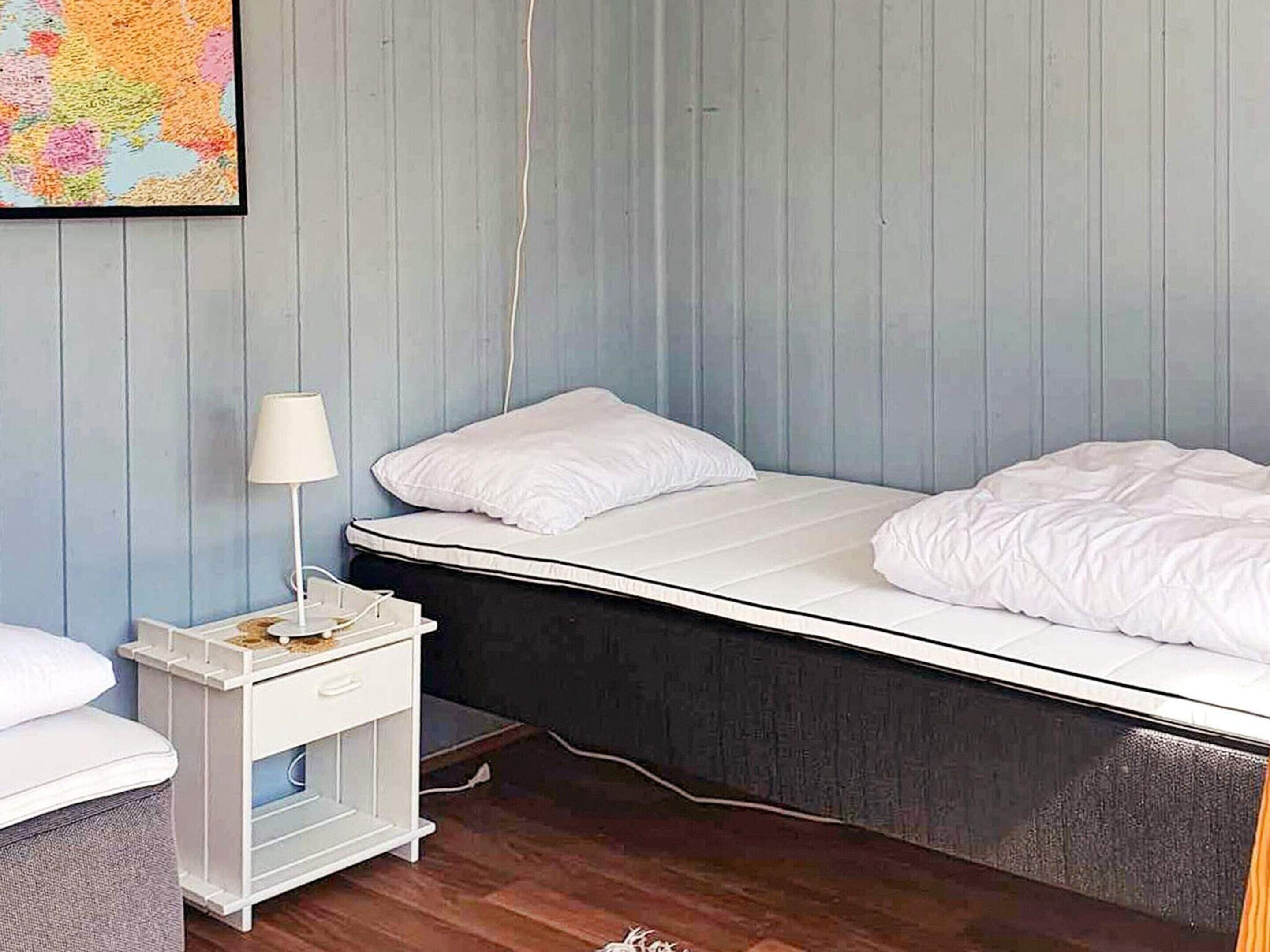 A small, single-bed room with a white nightstand, a lamp, and a map on the wall.