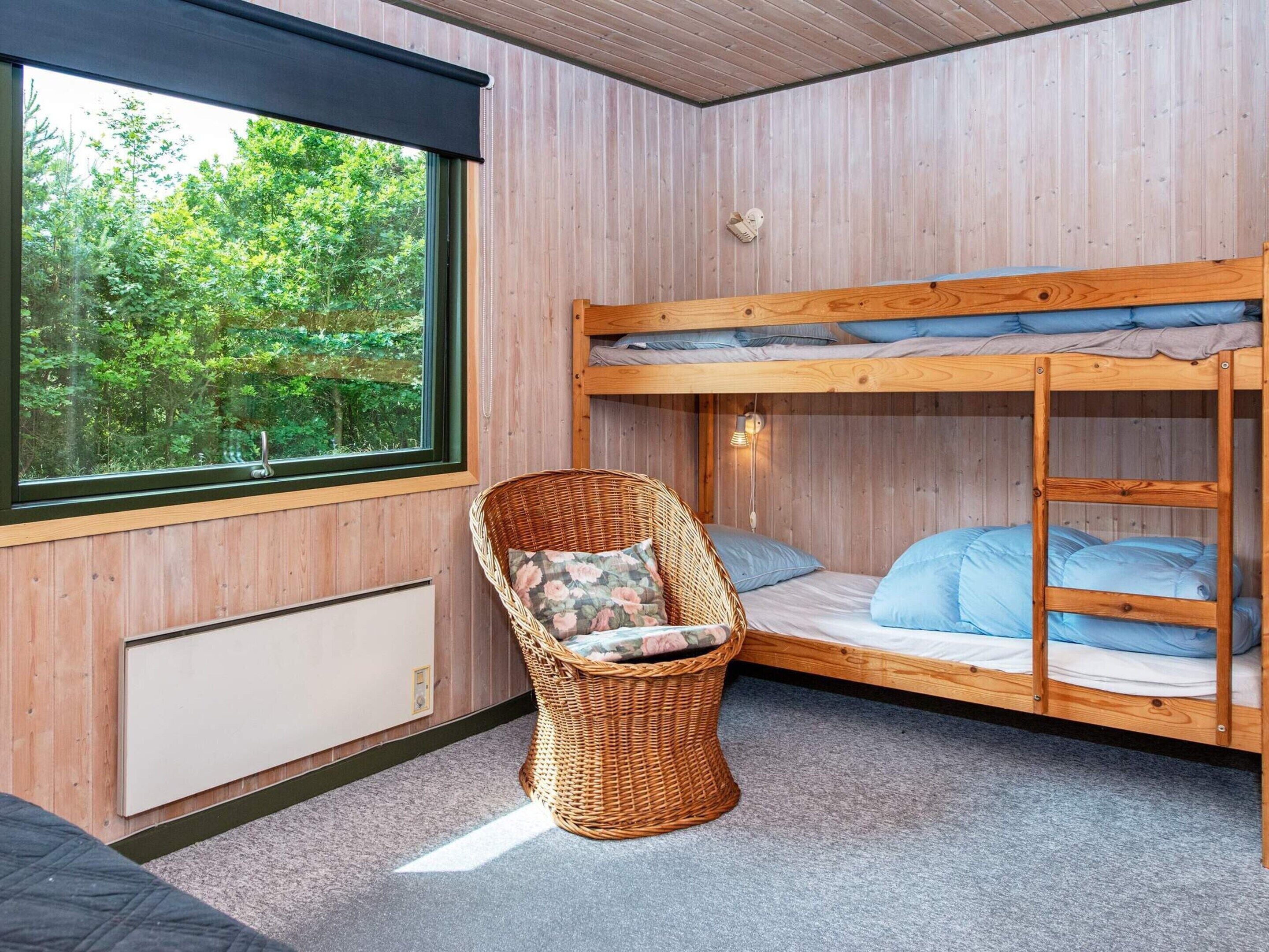 Una cama litera con vistas al follaje exterior, una silla de mimbre con cojín y una lámpara fijada a la pared.