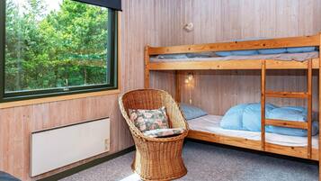 Una cama litera con vistas al follaje exterior, una silla de mimbre con cojín y una lámpara fijada a la pared.