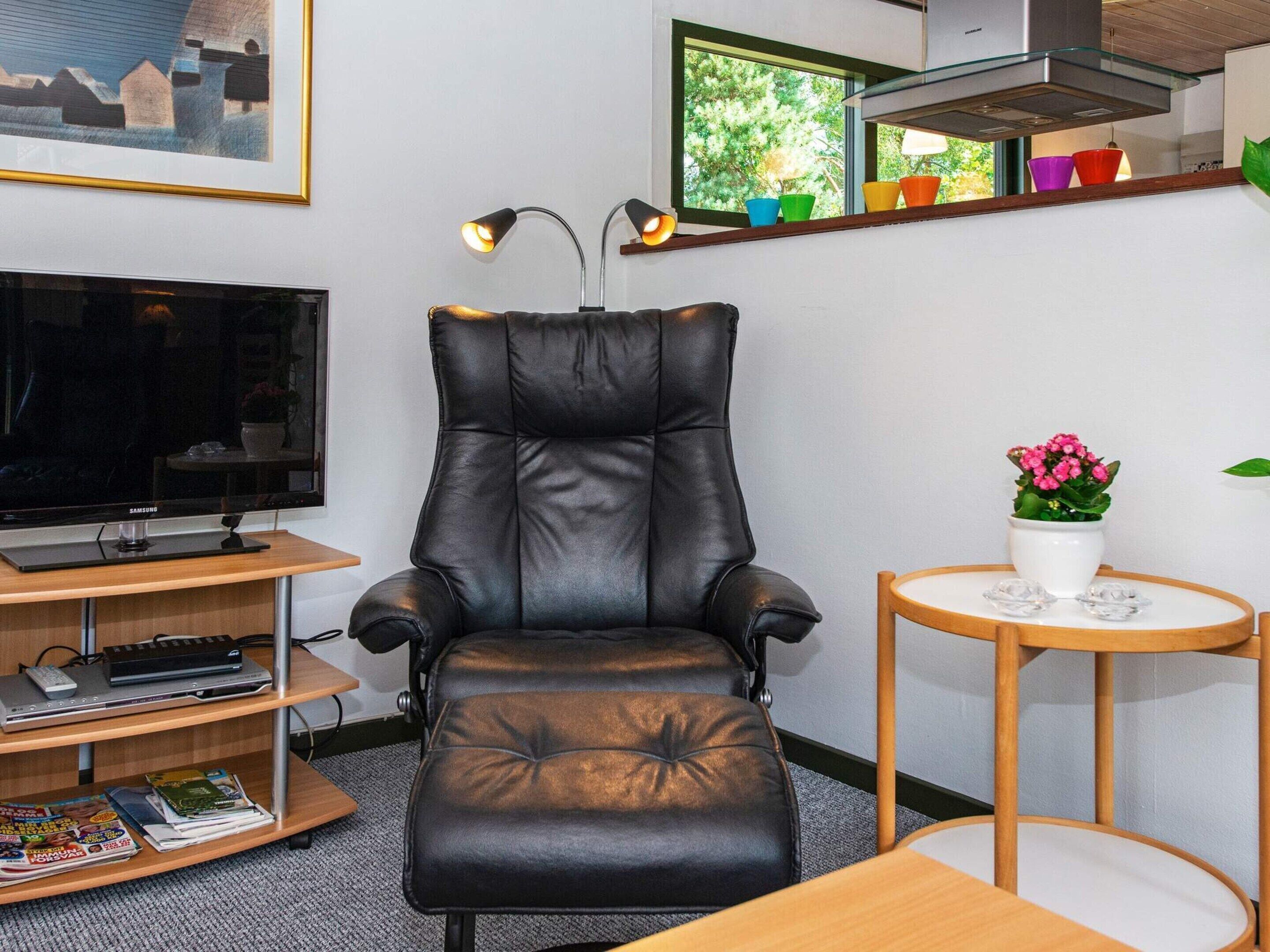 A modern living room with a leather recliner, a flat-screen TV, a small table with a potted plant, and a shelf with colorful cups.