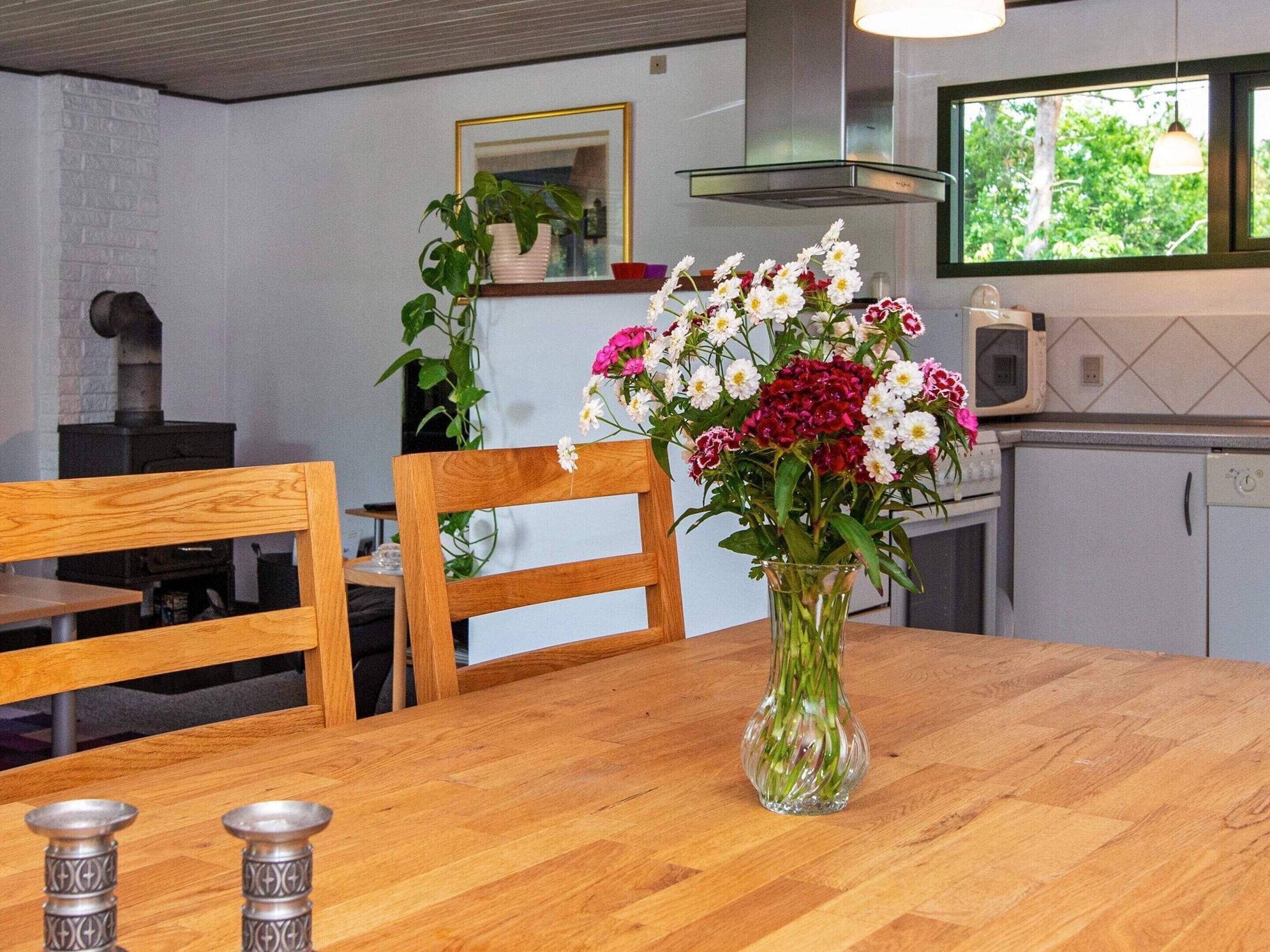 Et træbord med blomster i en vase, stole, en komfur og et vindue med udsigt til træer.