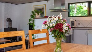 Une table de salle à manger en bois, ornée d’un vase de fleurs, entourée de chaises, avec un poêle à proximité et une fenêtre donnant sur des arbres.