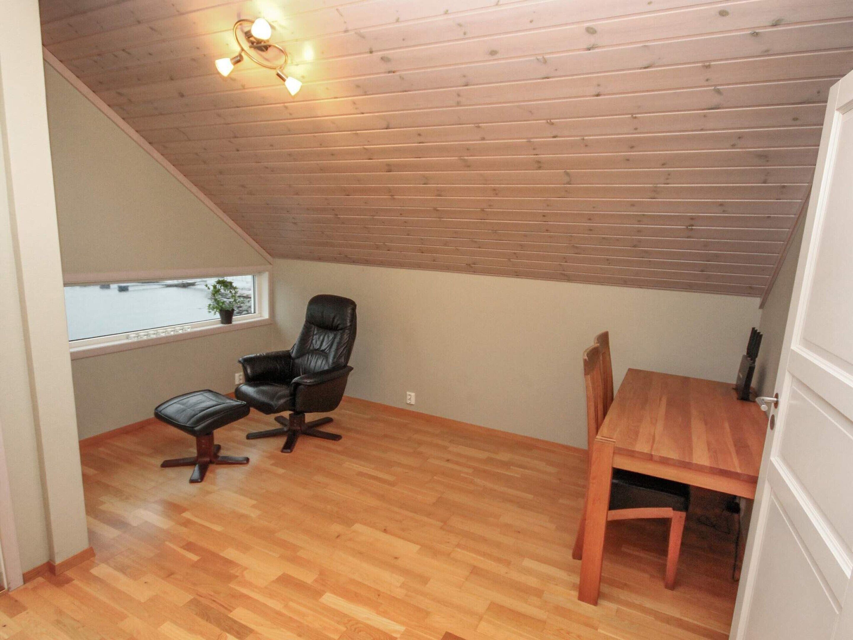 A small, sloped-roof room with a wooden floor, a black leather chair, a wooden dining table, and a window with a view of the outside.