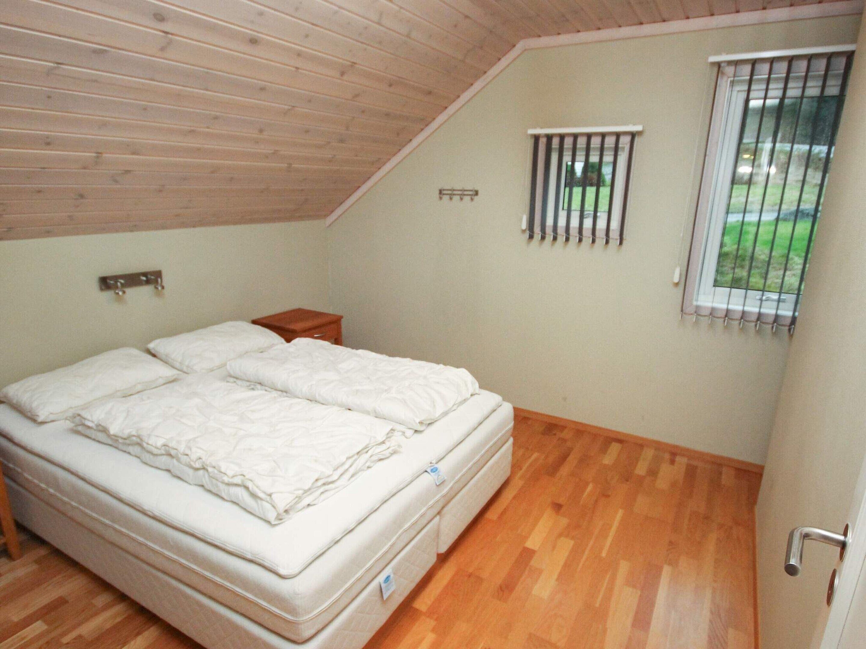 A bedroom with a wooden ceiling, a single bed, a small bedside table, a window with bars, and a view of greenery outside.