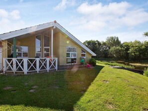 Ett trähus med en veranda och en liten terrass, omgivet av en gräsklädd trädgård och en damm.