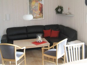 Una sala de estar con un sofá negro, dos sillas de madera, una mesita con un candelabro de vidrio y un estante en la pared con adornos.