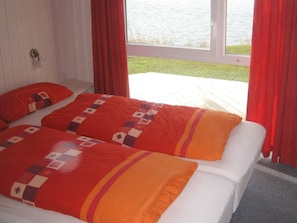 A double bed with a patterned bedspread and pillows, a window with red curtains, and a view of a body of water.