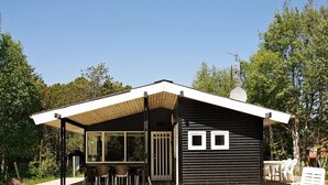 Ett modernt utomhusområde med plats för måltider, bestående av ett bord och stolar, en överbyggd sittplats och en byggnad med ett sluttande tak och två fönster.