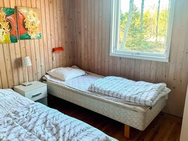 Una habitación pequeña y acogedora con paredes de madera, una cama individual, una mesita de noche, una lámpara y una ventana con vistas a árboles.