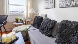 A modern living room with a grey sofa, fluffy grey cushions, a small round table with a lamp, and a window with curtains.