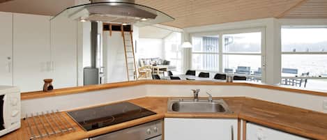 A modern kitchen with a stove, oven, and sink, wooden countertops, and a view of the sea through large windows.