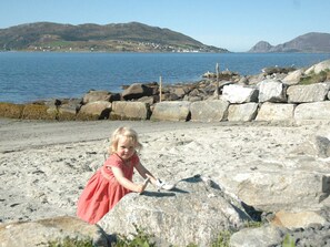 Uma criança brincando em uma praia rochosa, com vista para o mar e colinas ao fundo.