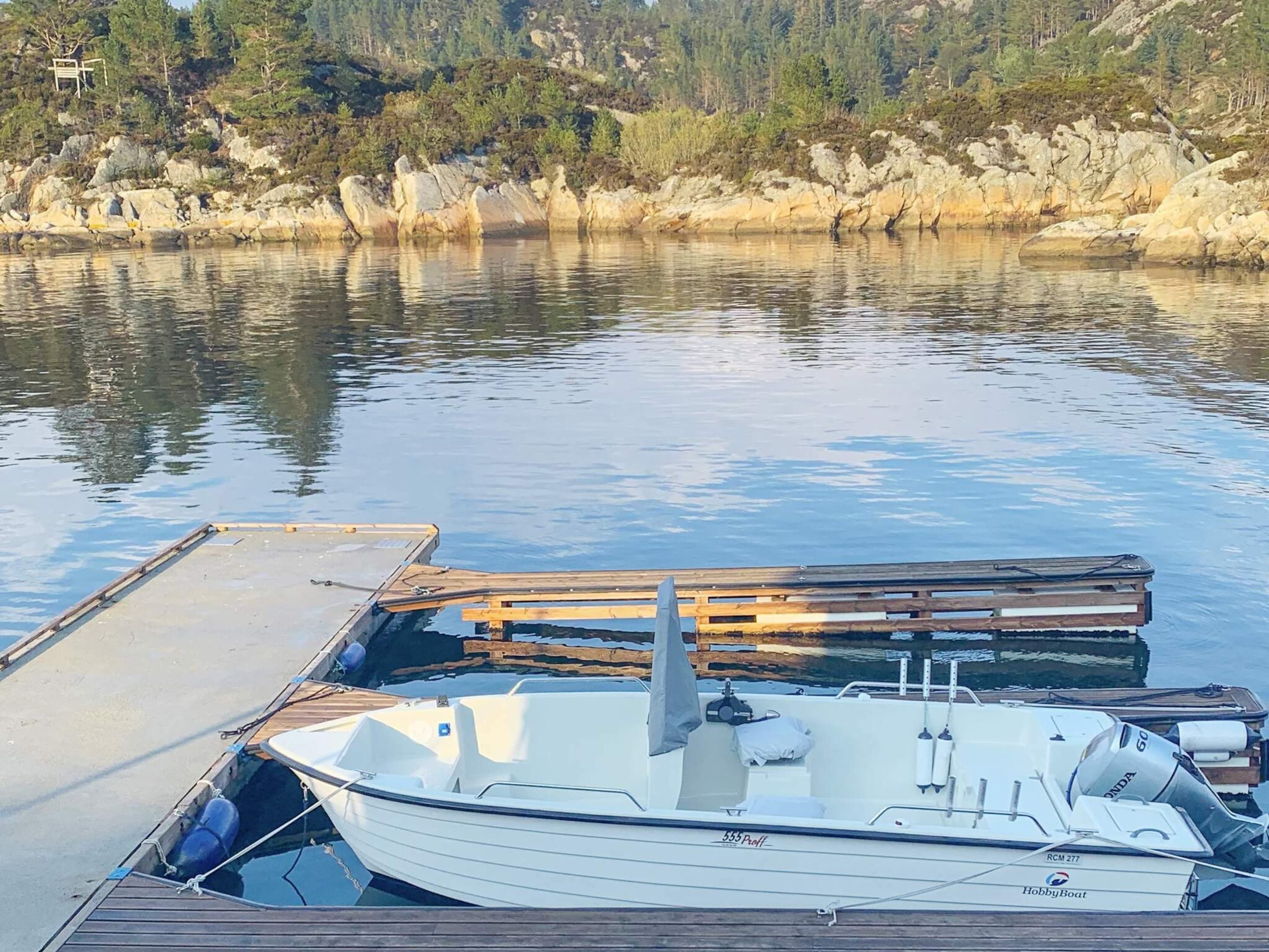 A boat docked at a wooden pier with a rocky shoreline and trees in the background.
