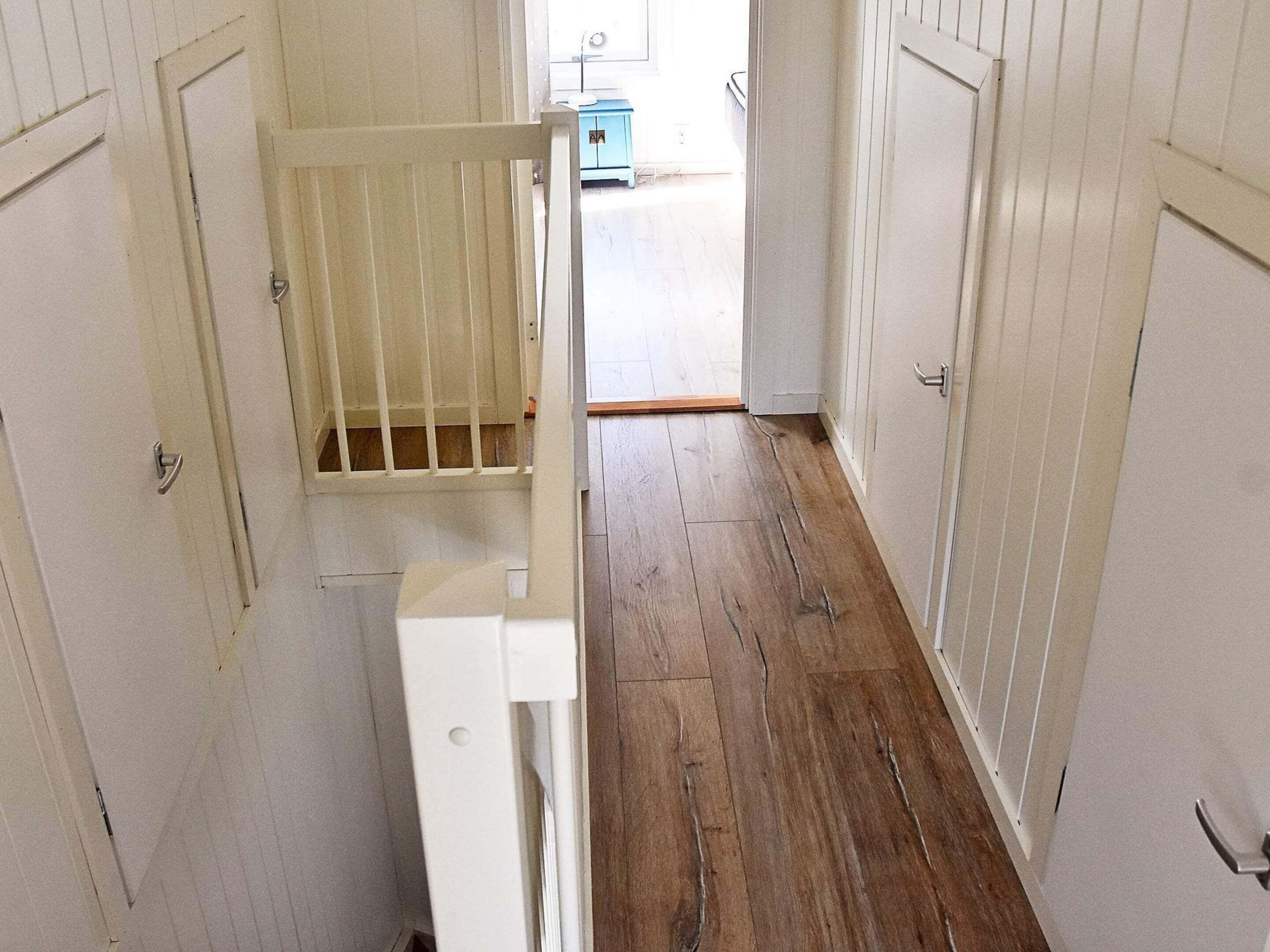 Un couloir étroit avec un plancher en bois, des murs blancs et une barrière de sécurité pour bébé au milieu.