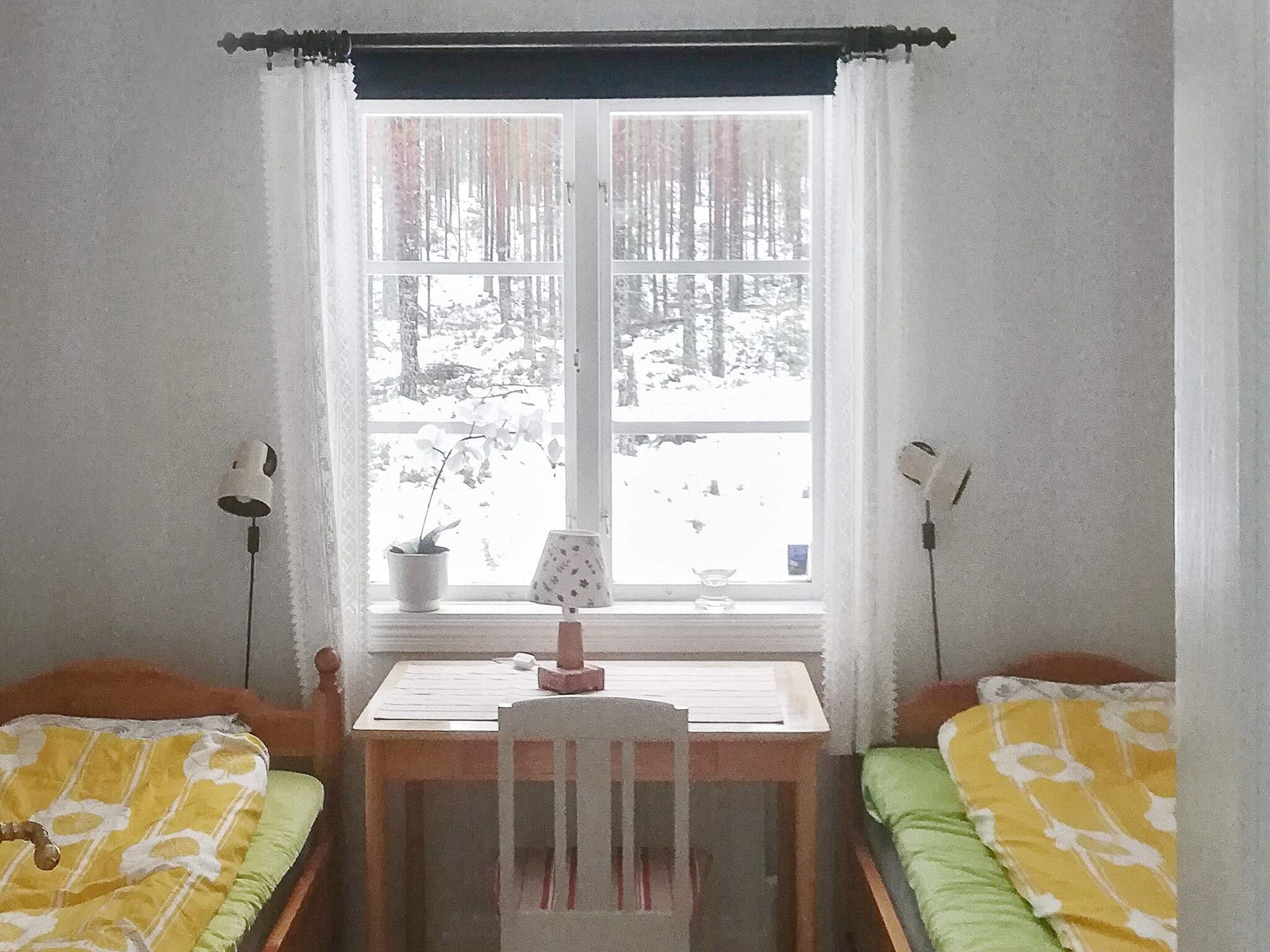 A room with two single beds, a desk, a chair, a window with a view of a snowy landscape, and a lamp on the desk.