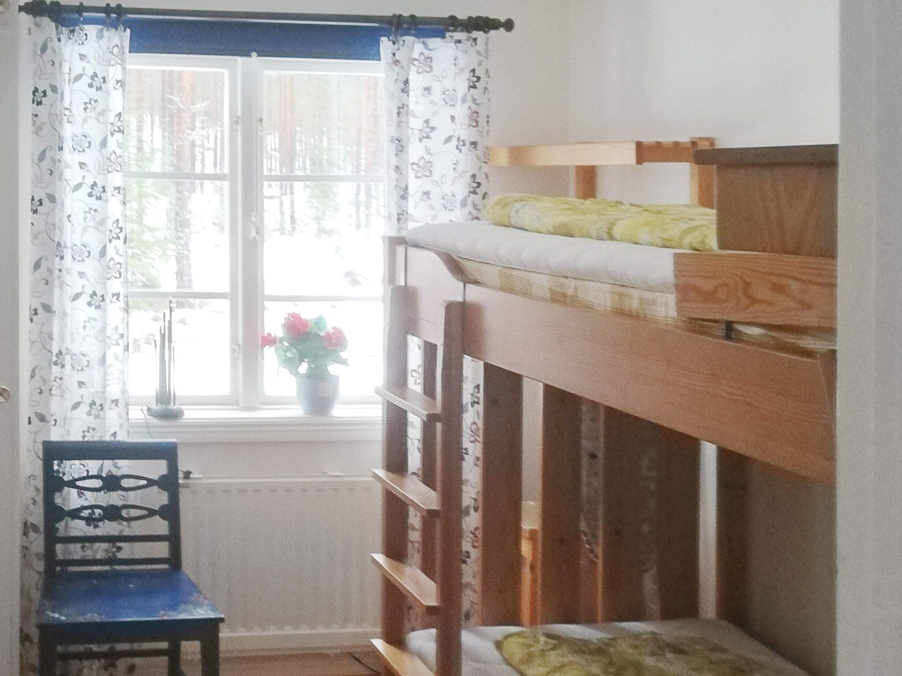 A bunk bed with a wooden frame and a mattress, a chair with a floral pattern, and a window with curtains.