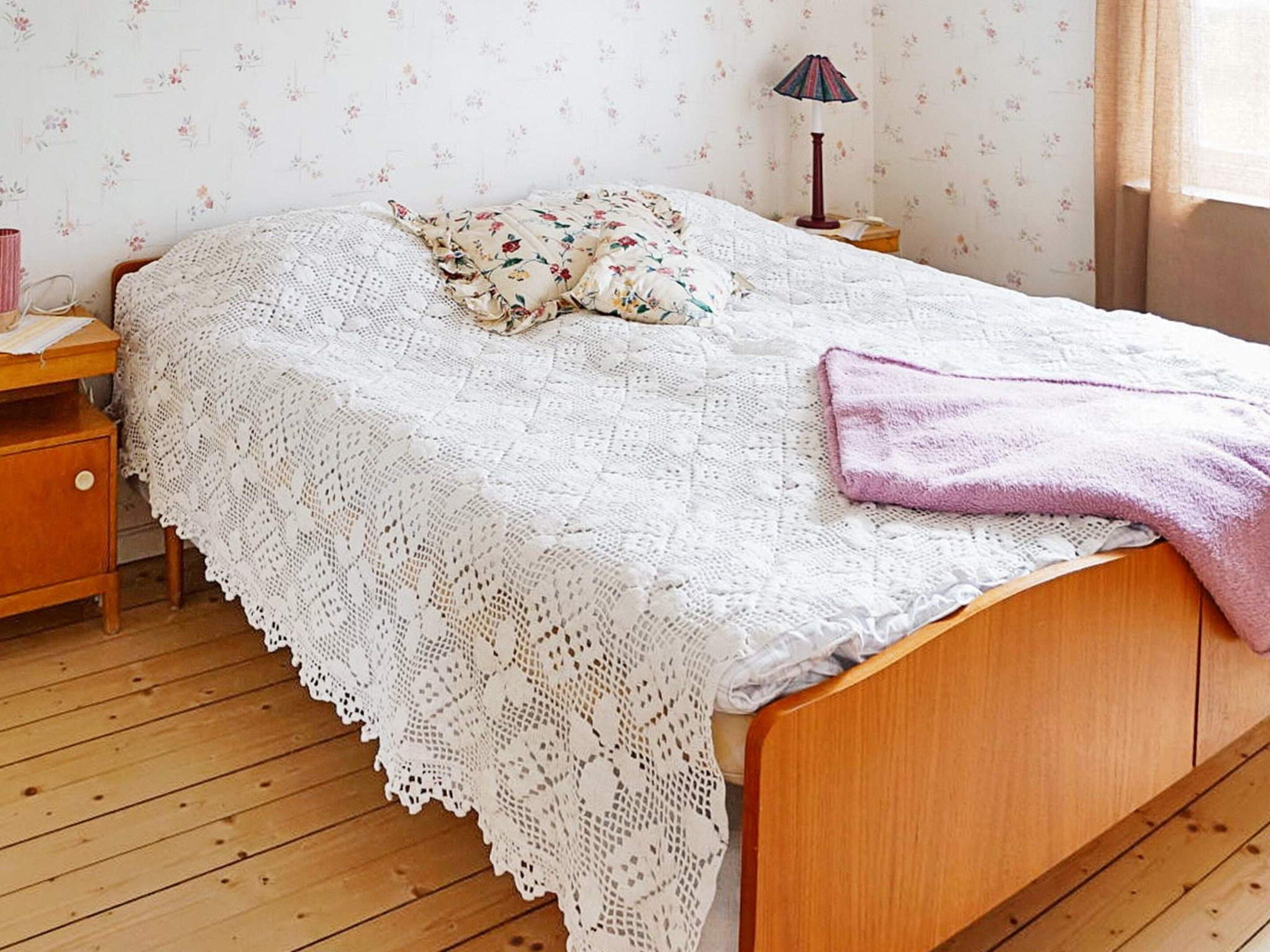 A bedroom with a wooden bed, a white lace bedspread, a floral pillow, a pink towel, and a nightstand with a lamp.