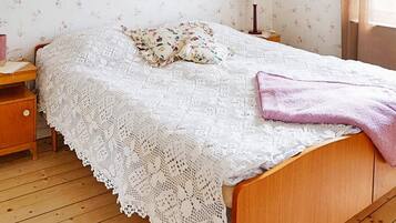 A bedroom with a wooden bed, a white lace bedspread, a floral pillow, a pink towel, and a nightstand with a lamp.