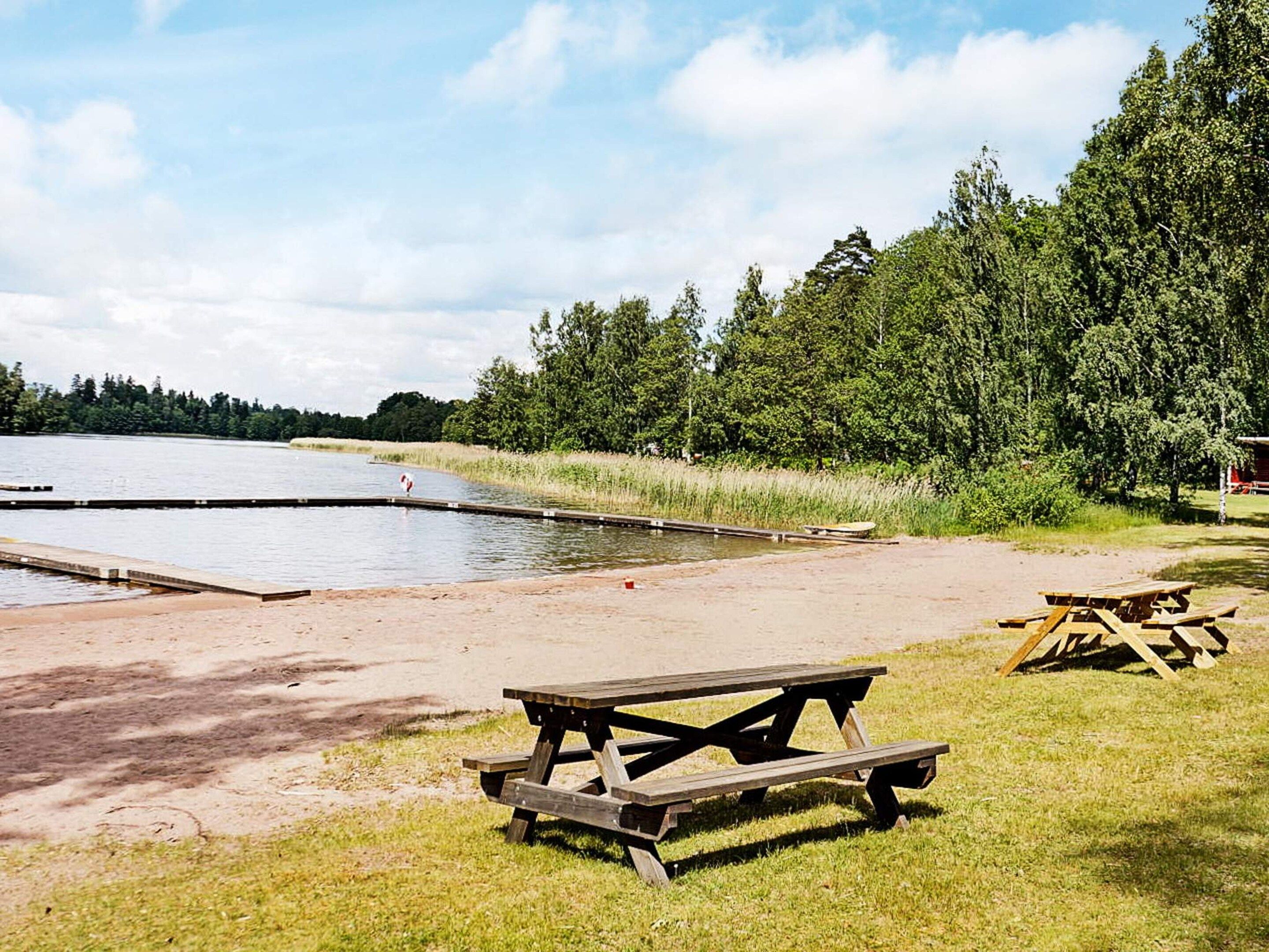 Un área de picnic junto al lago con banco de madera y mesas, rodeada de árboles y un cielo despejado.