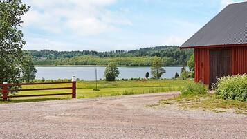 A gravel driveway leading to a red barn with a metal roof, surrounded by greenery and a lake in the background.